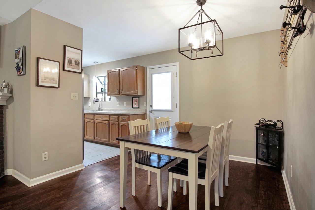 Dining room with a wooden table and four white chairs. Overhead, a modern chandelier hangs. Adjacent kitchen features wooden cabinets and a window.