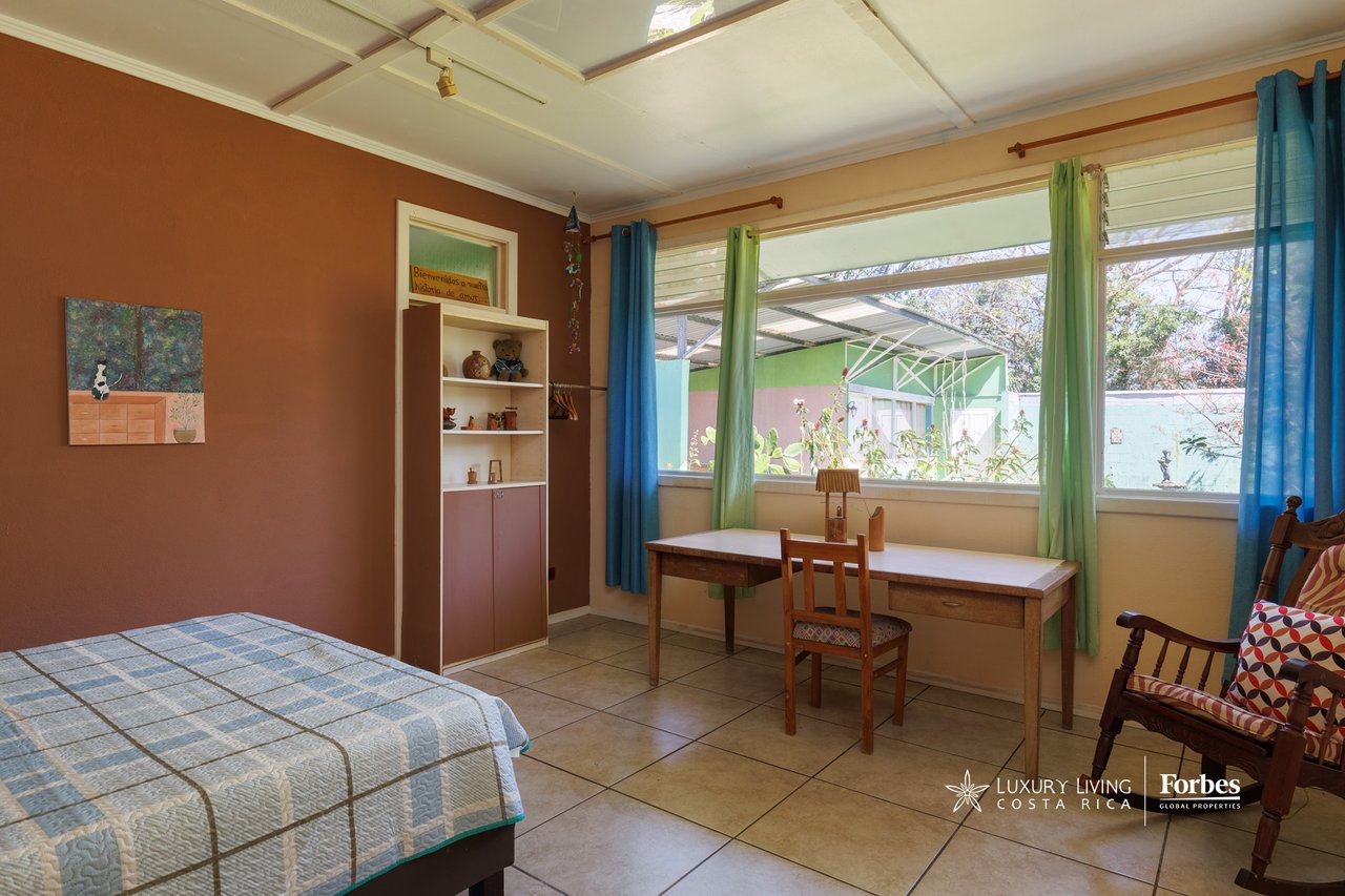 Furnished bedroom with a blue-and-white quilt, wooden desk, and colorful curtains framing wide windows.