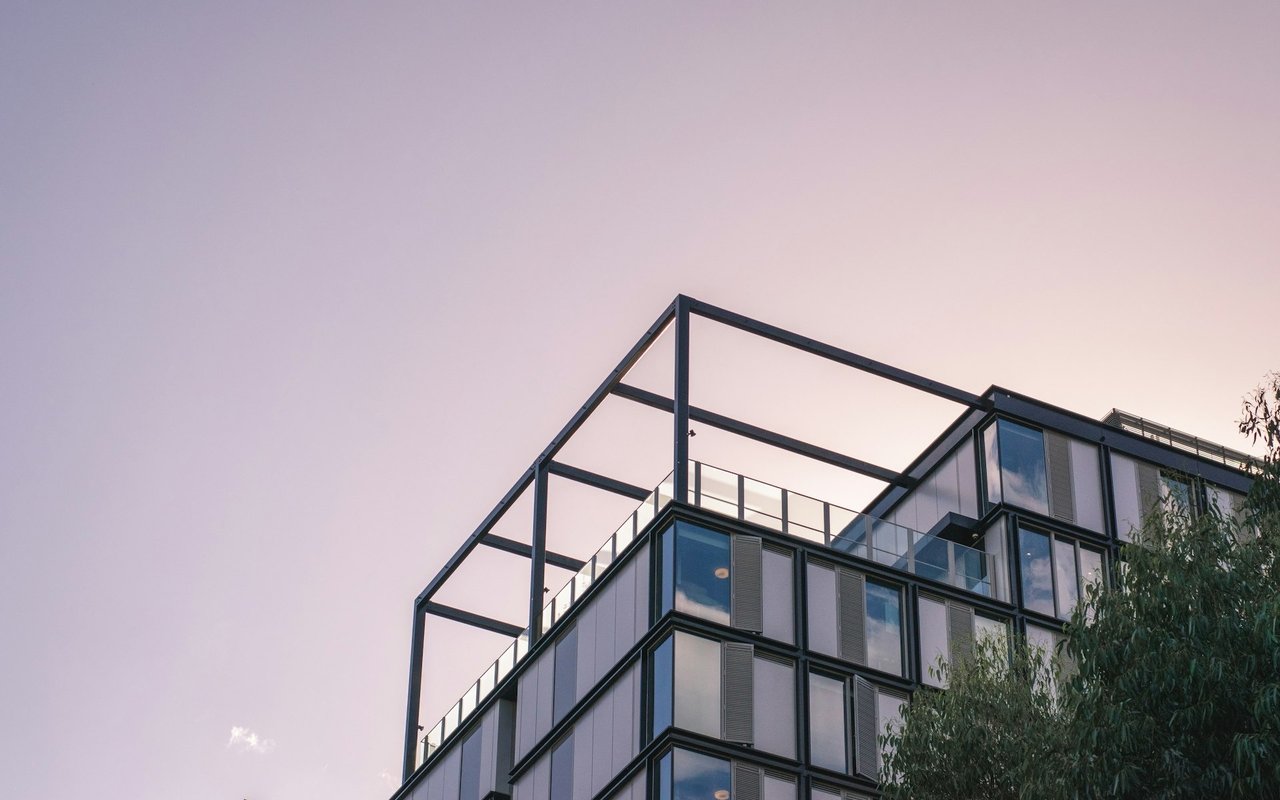 Modern glass-and-metal building with an open steel rooftop frame against a pink dusk sky.