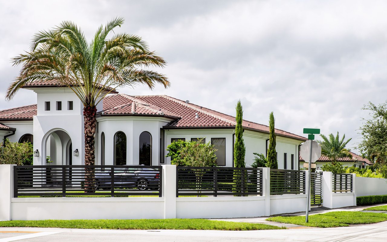 White stucco home with a terracotta tile roof, arched entry, and black fence behind palm trees.