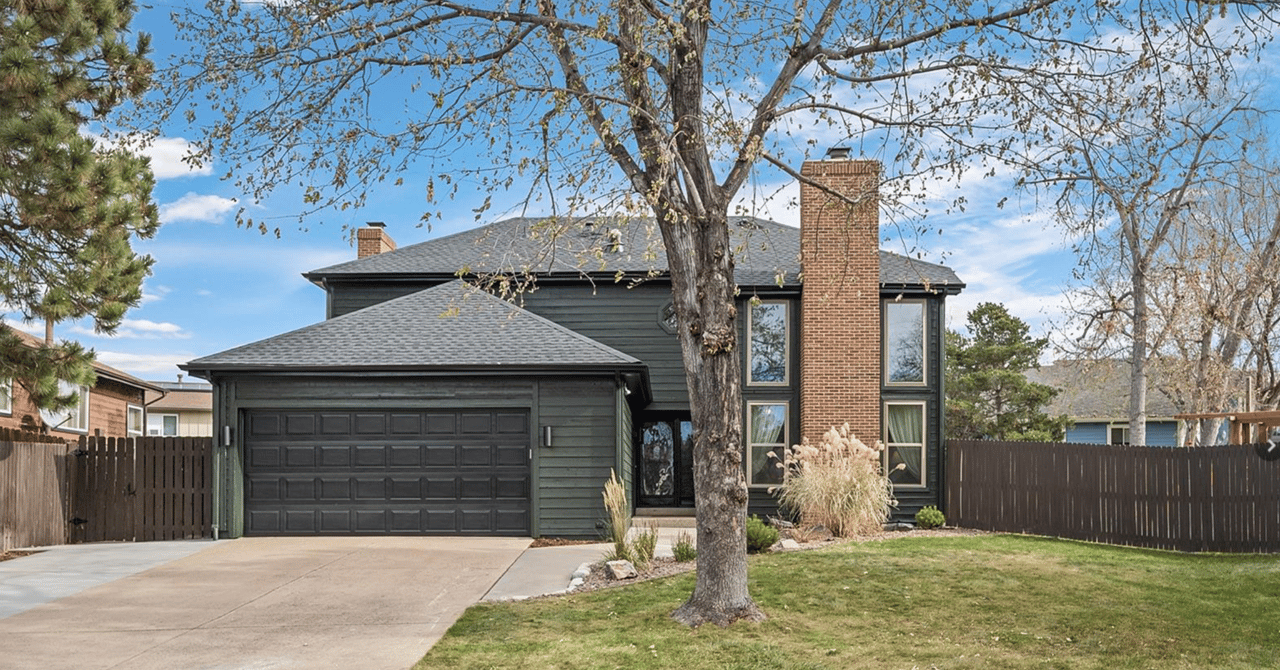 Two-story dark green home with a double garage, brick chimney, mature front-yard tree, and fenced lawn.