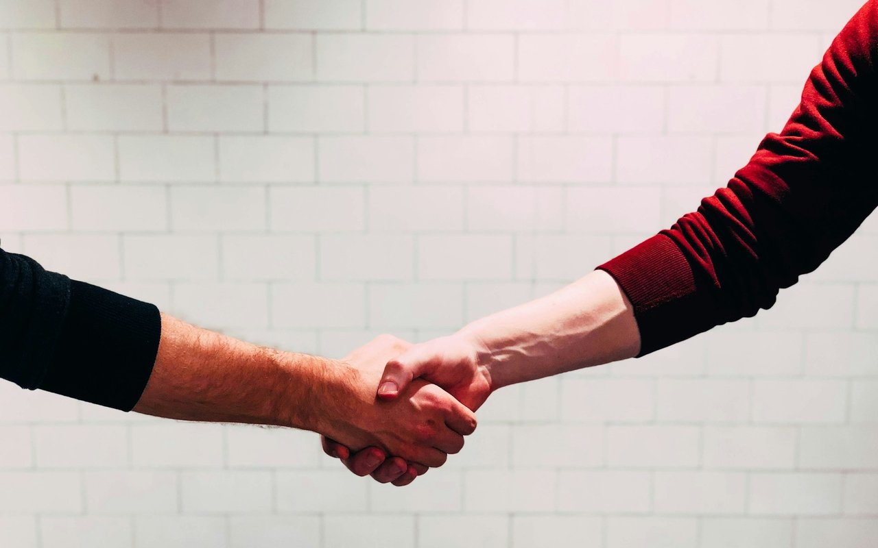 Two people shake hands in front of a pale block wall.