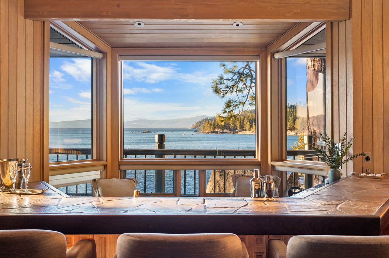 Wood-paneled kitchen bar with wide windows overlooking a lake and forested shoreline.