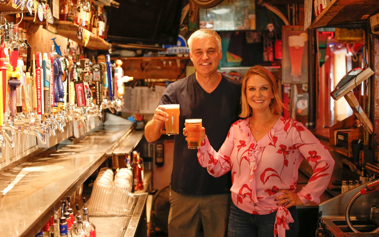 A man and woman smiling and holding glasses of beer in a rustic bar, with beer taps and bottles visible in the background.