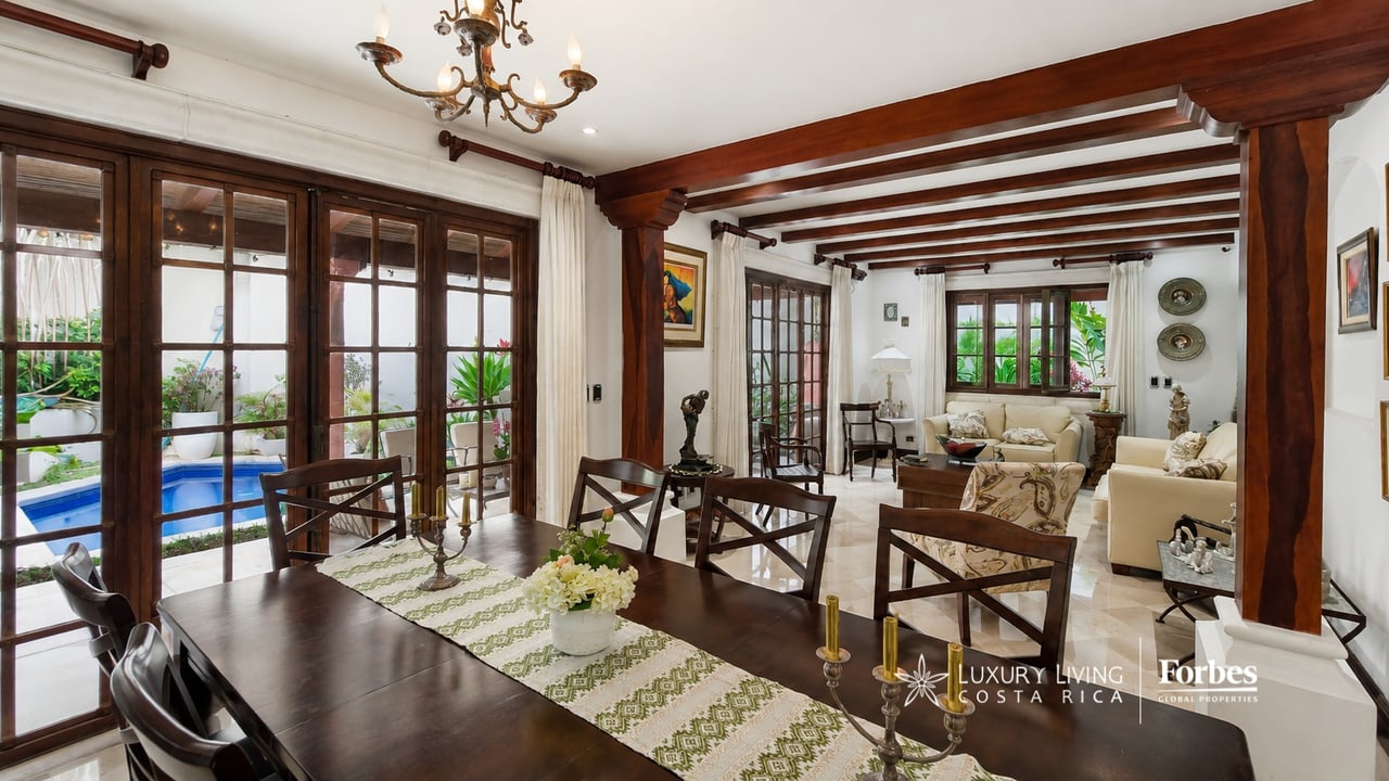 Dining and living room with dark wood beams and glass doors overlooking a small swimming pool.