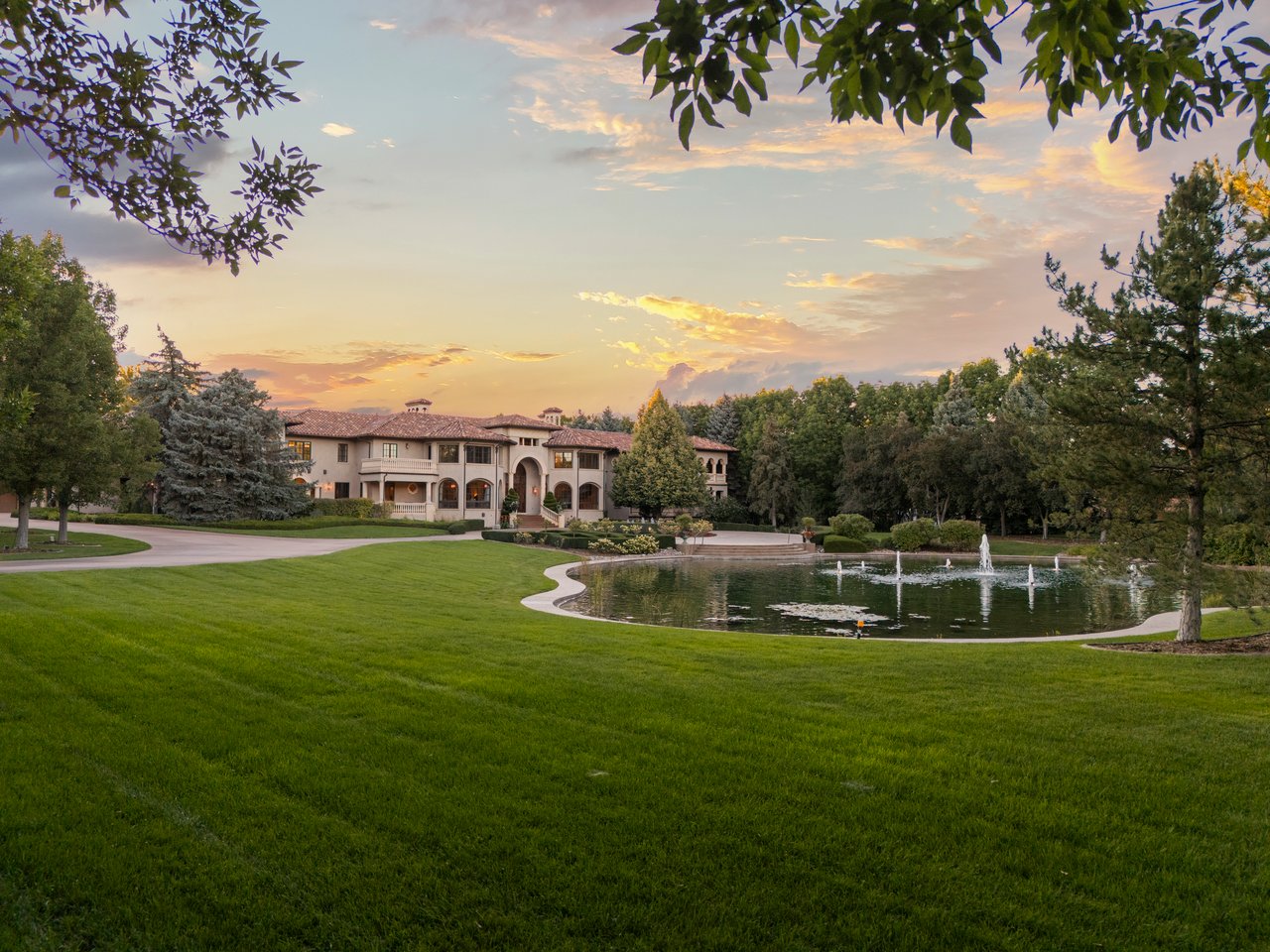 Estate grounds with a fountain pond, sweeping lawn, and mature trees at dusk.