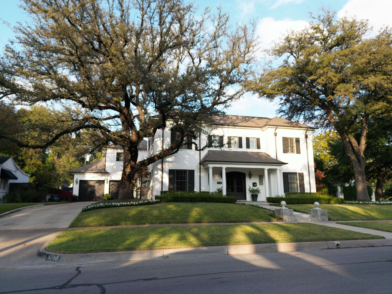 White two-story house with a columned porch, attached garage, and mature trees framing the front yard.