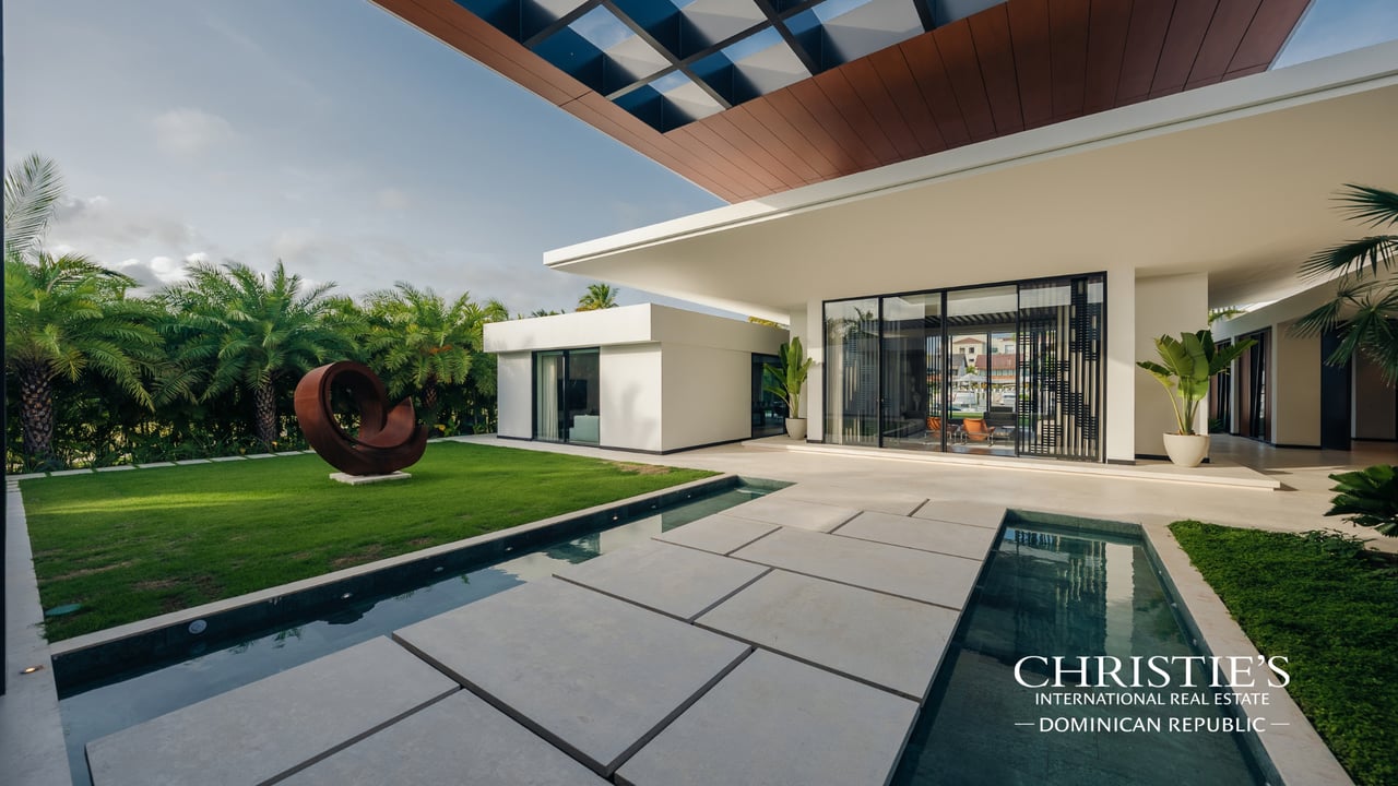 Modern villa with a long reflecting pool, stone terrace, tropical palms, and abstract lawn sculpture.