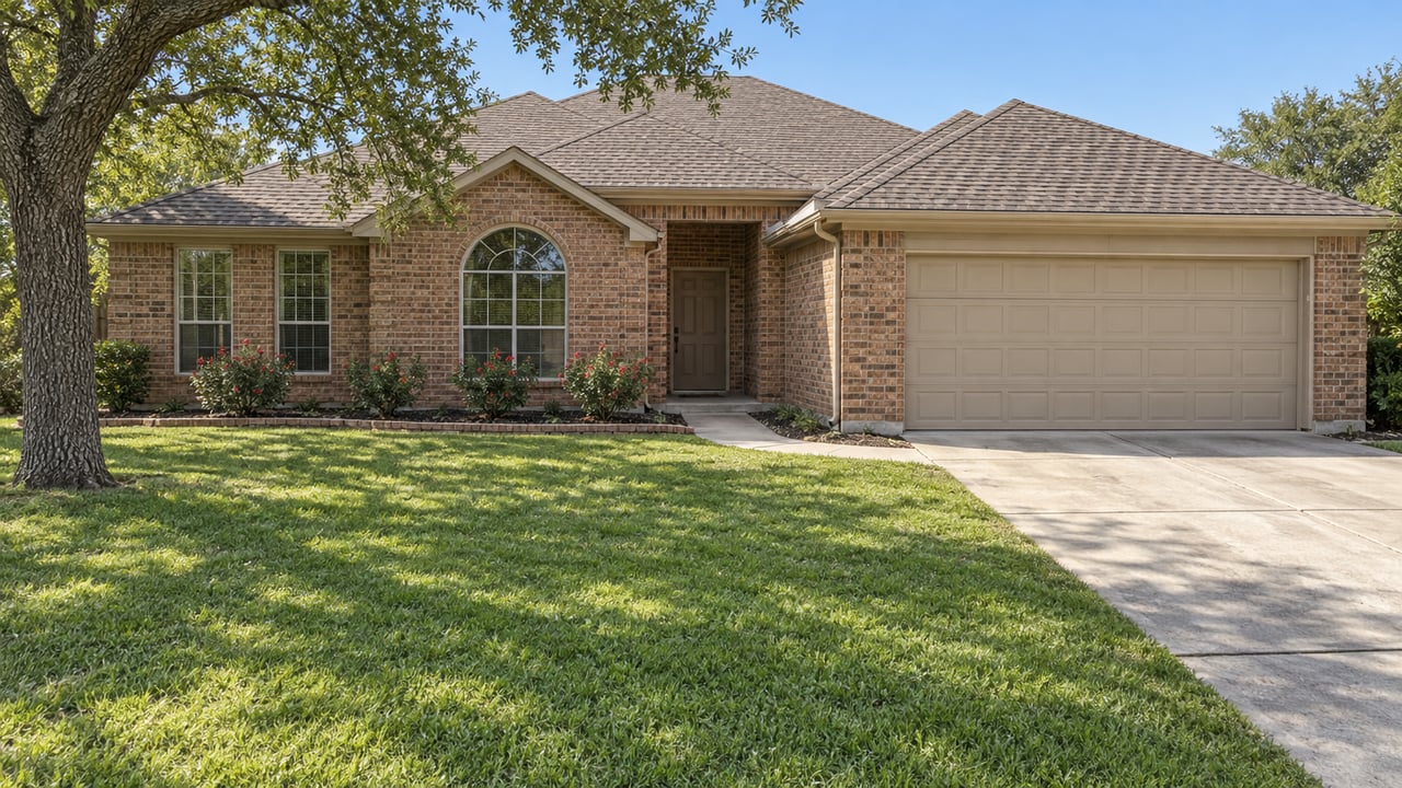 Wide view of a single-story russet brick home with gabled shingle roofs, a two-car garage, deep green lawn and a mature post oak.