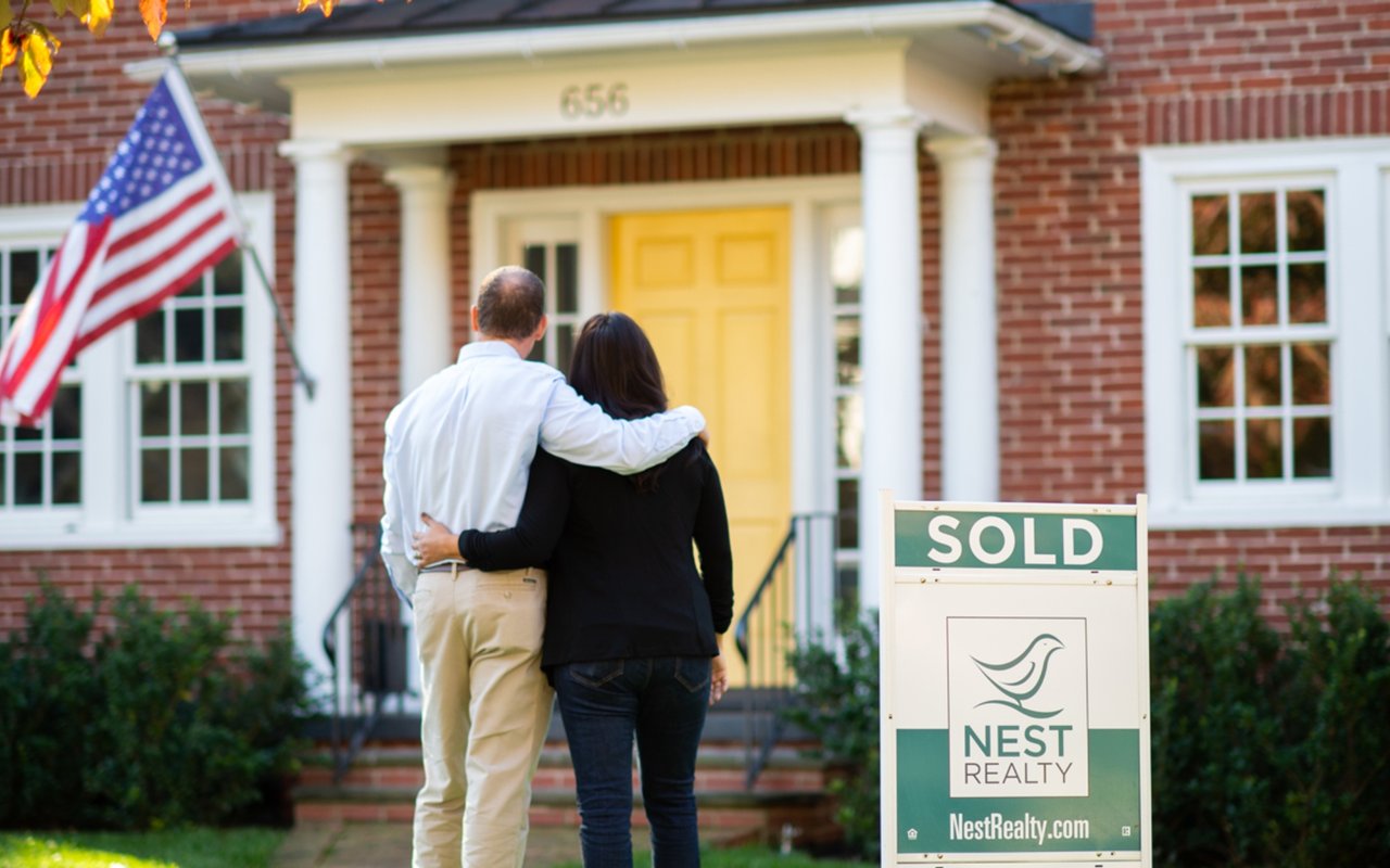Brick house with a yellow door, a SOLD sign, and a couple standing in the front yard.