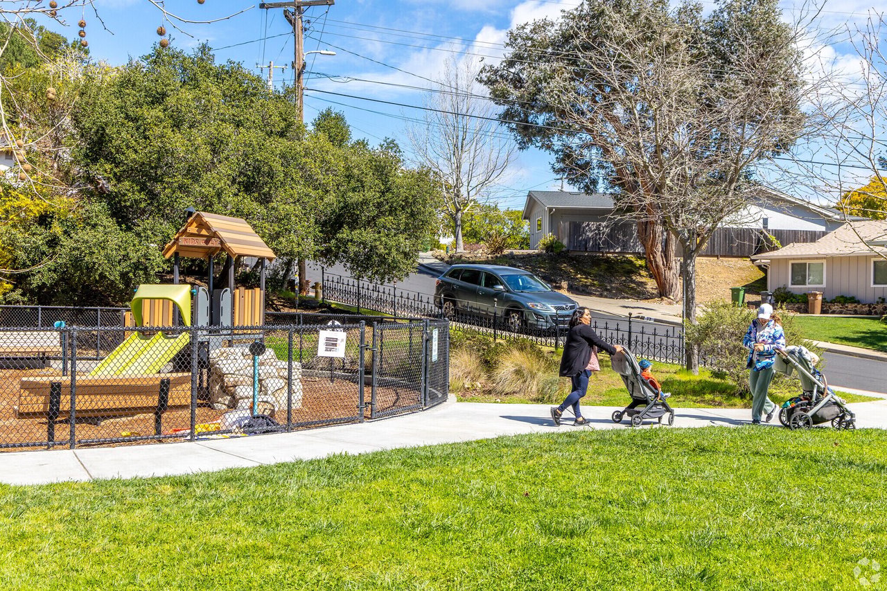 Neighborhood playground beside a sidewalk, with two adults walking strollers and homes across the street.