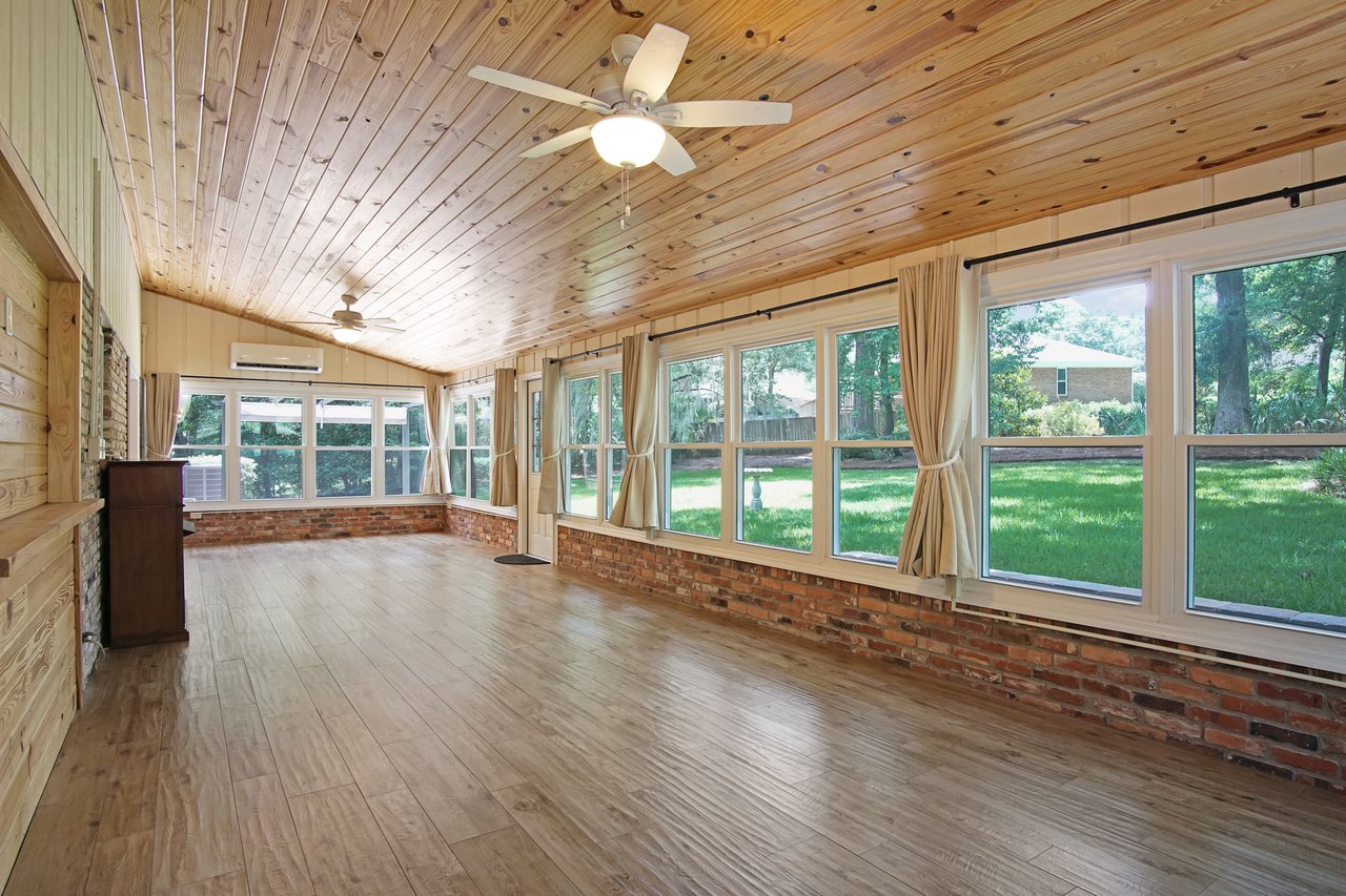 Bright sunroom with wooden floors and ceiling, large windows with beige curtains, and brick accents. Overlooks a green backyard. Airy and serene.