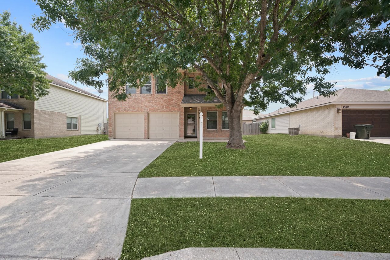 Two-story brick house with a double garage, wide driveway, and large tree shading the front lawn.