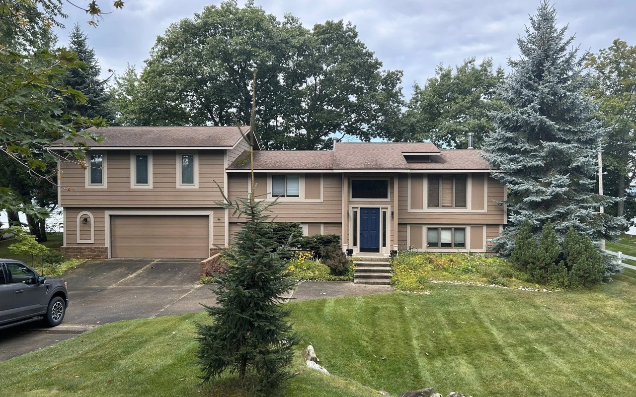 Tan split-level house with a blue front door, attached garage, and mature trees.