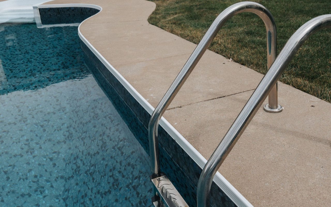 Swimming pool with a stainless-steel ladder beside a light stone deck and dry grass.