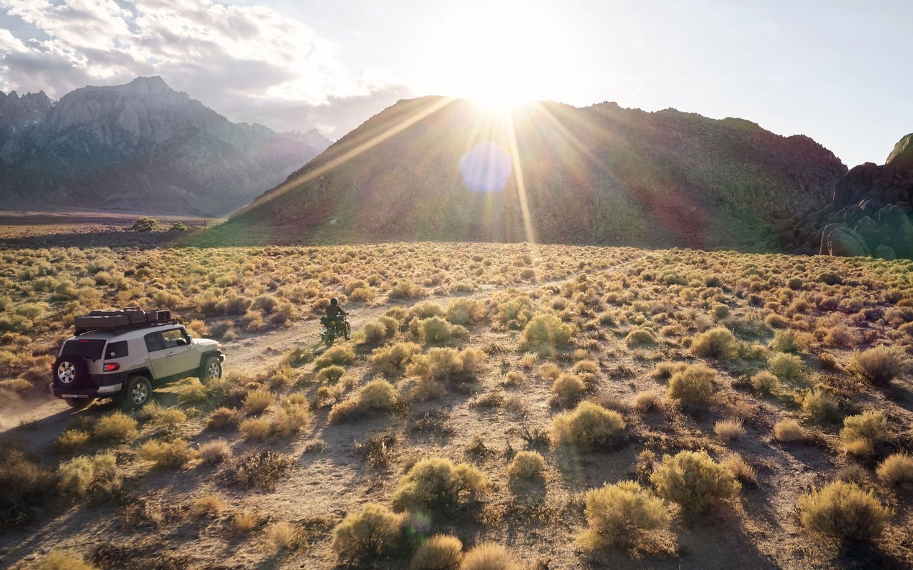 Desert mountains at sunset with an off-road SUV and motorcyclist crossing scrubland.