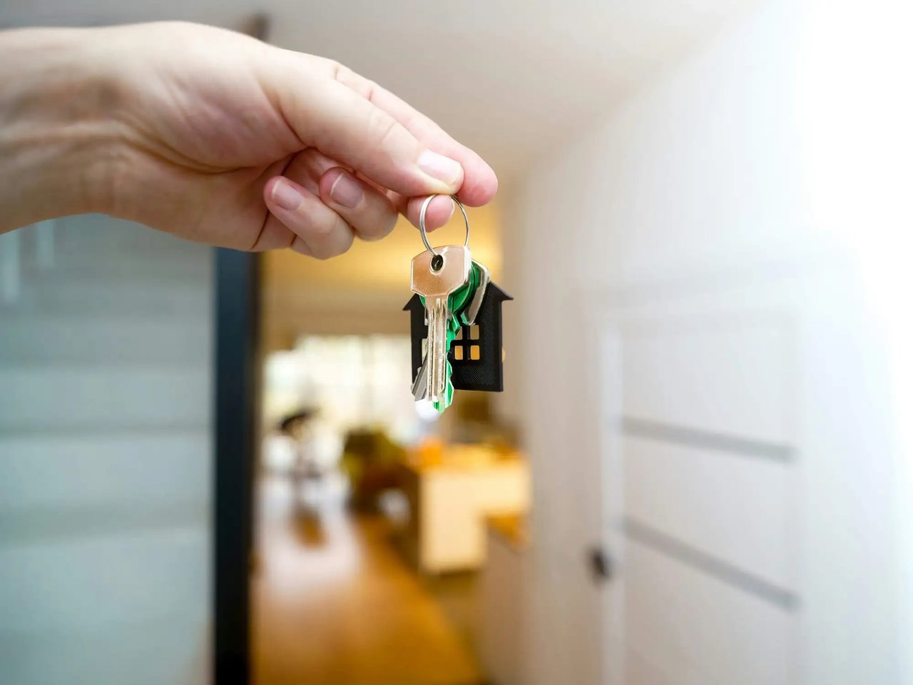 A hand holds house keys with a small house-shaped keychain in a blurred home interior.