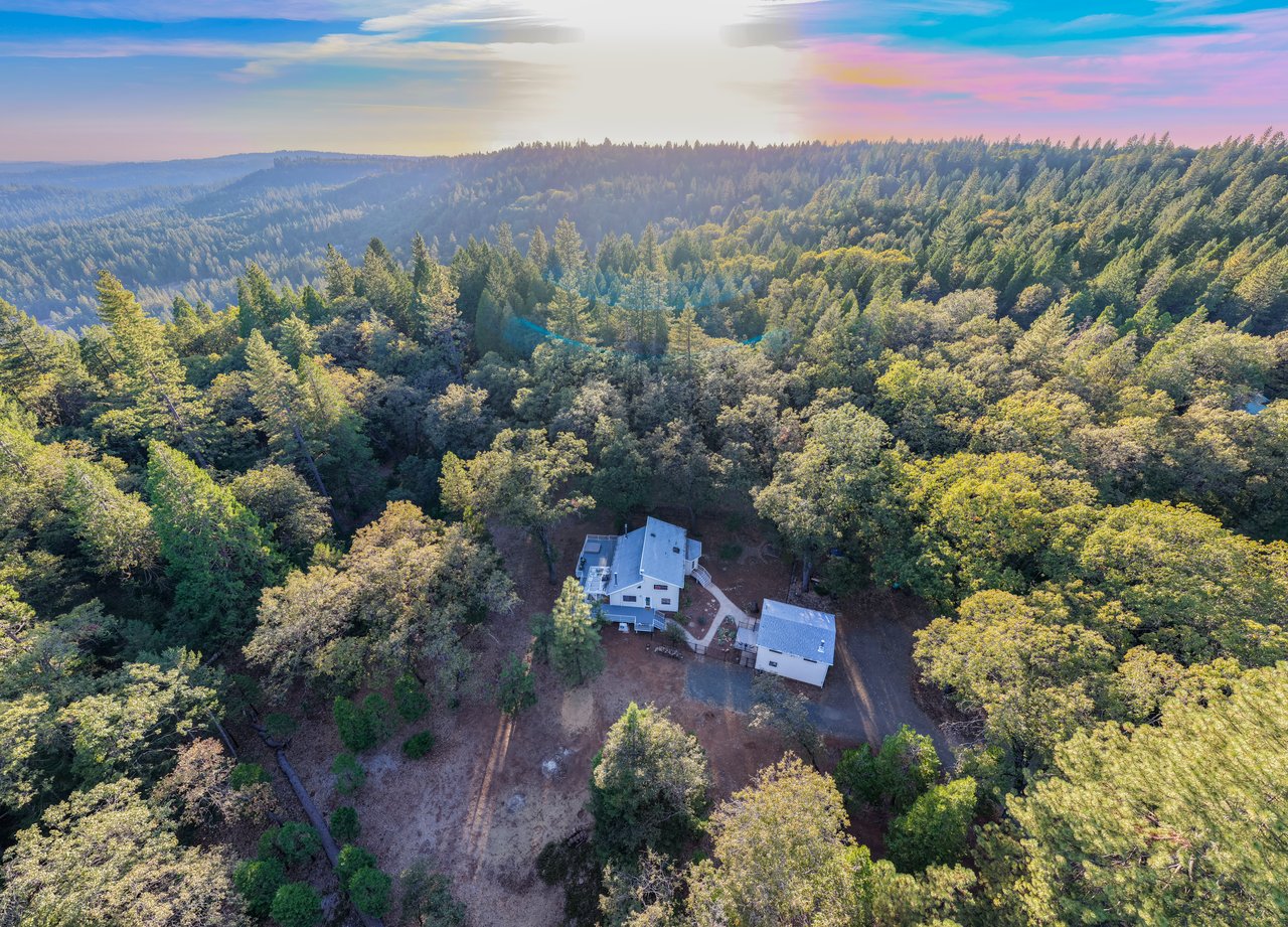 Aerial view of a home and detached outbuilding amid dense forest and rolling wooded hills.