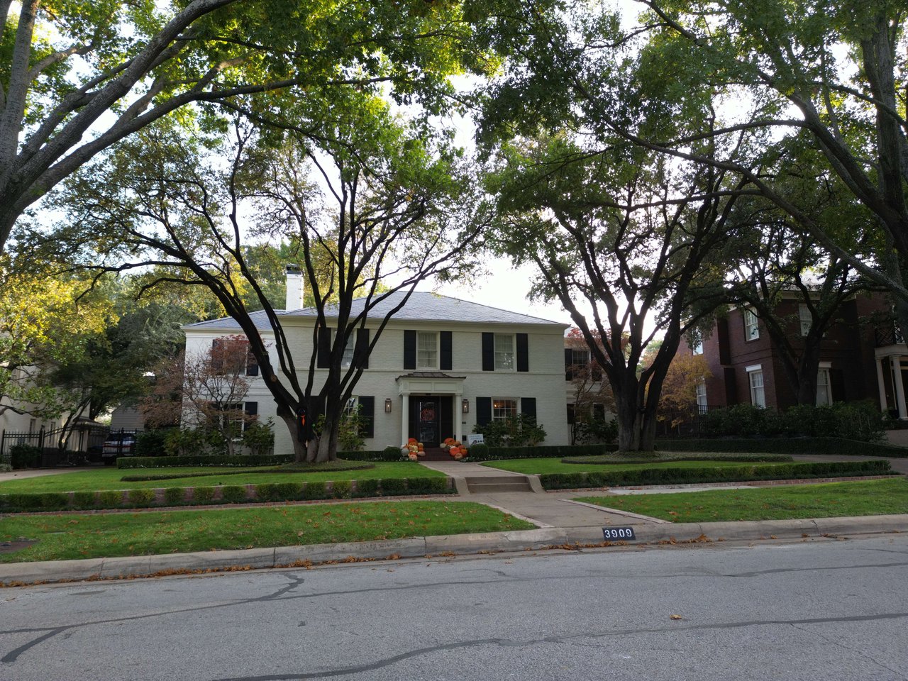 White two-story brick home framed by mature trees, with pumpkins on the front steps.