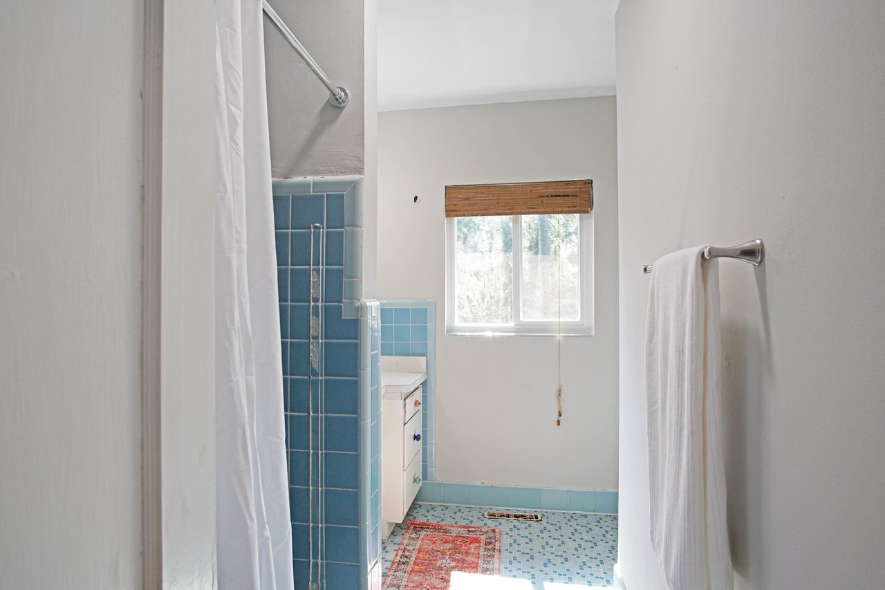 Bright bathroom with blue tiles and a white shower curtain on the left. A window with bamboo blinds illuminates the space. A towel hangs on the right.