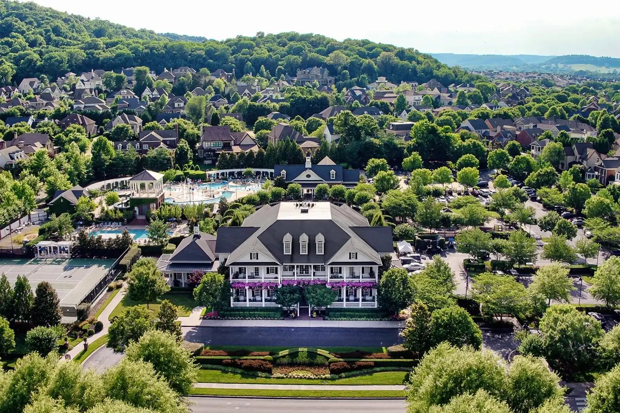 Aerial view of a residential community clubhouse, pools, and tennis courts among tree-lined homes.