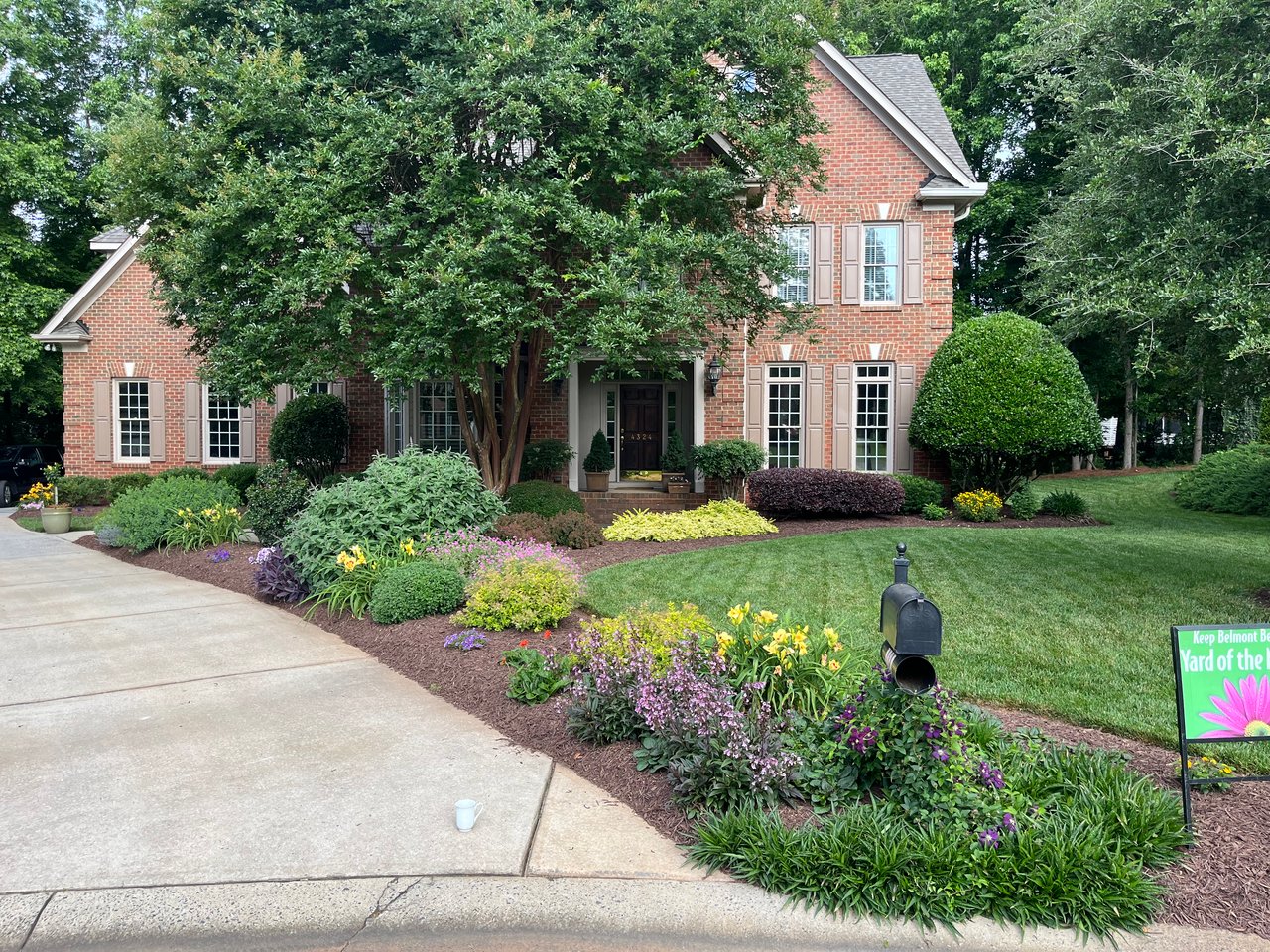 Red brick two-story home with a broad driveway, mature trees, and colorful landscaped flower beds.