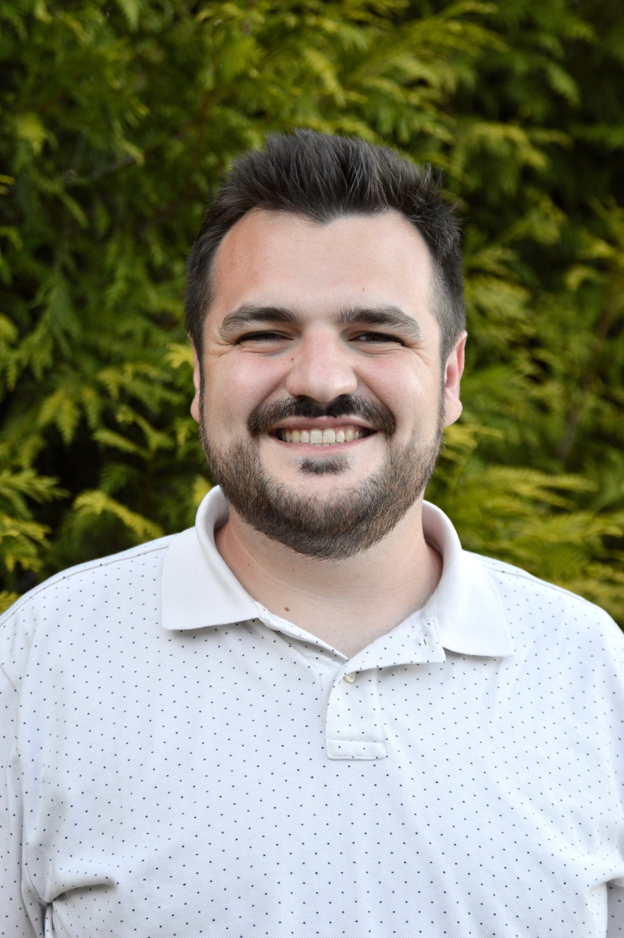 Trent Kelleher smiling in a white patterned polo against leafy greenery.