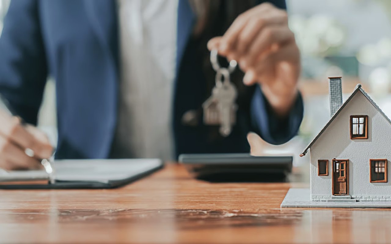 A person holds house keys while signing paperwork beside a model home.