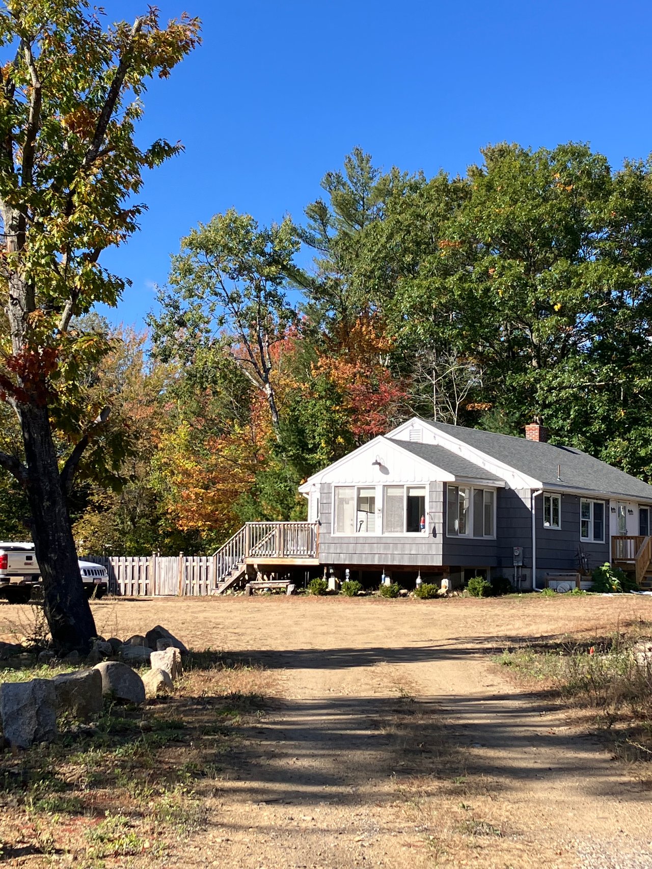 Gray single-story home with a raised deck, framed by autumn trees beside a dirt driveway.