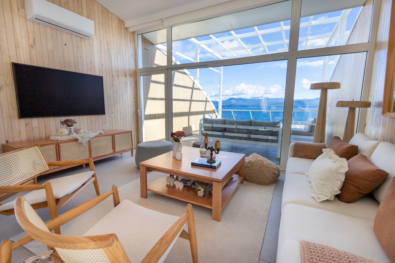 Living room with wood-paneled walls and wide windows overlooking water and mountains.