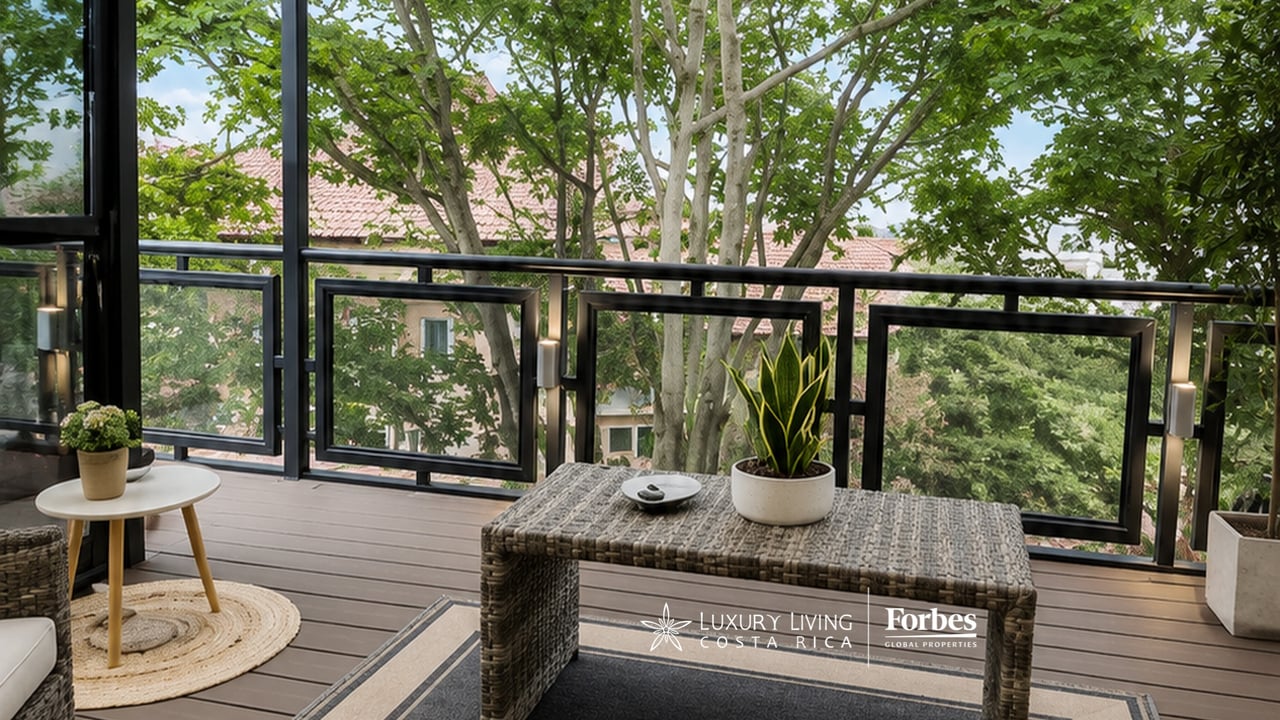 Furnished balcony with a woven table, potted plants, and black-framed glass railing overlooking leafy trees.