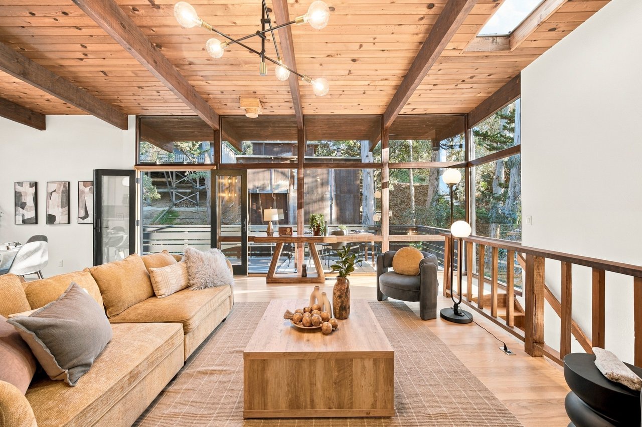 Living room with a vaulted wood ceiling, exposed beams, and floor-to-ceiling windows overlooking trees.