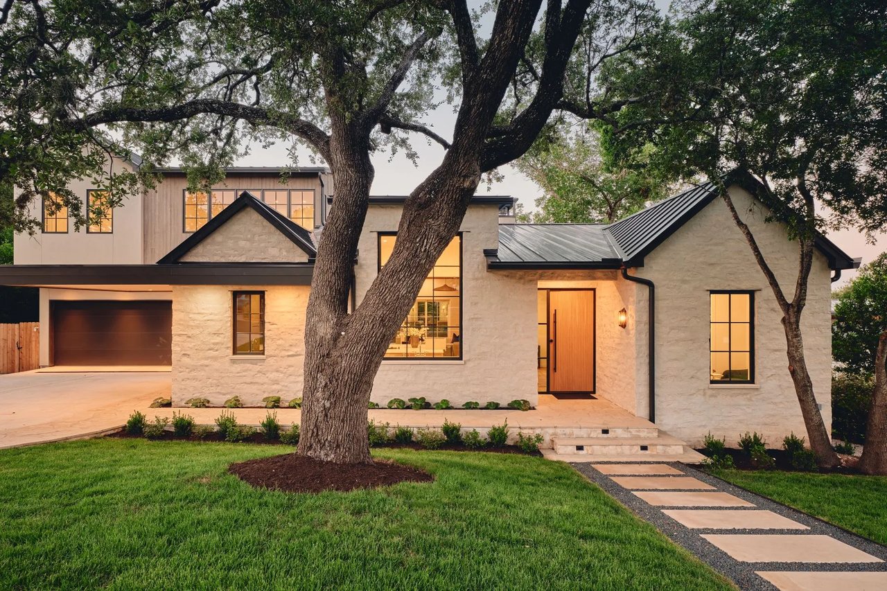 Modern stone-and-wood home with black metal roofs, warm-lit windows, and a mature oak tree in front.
