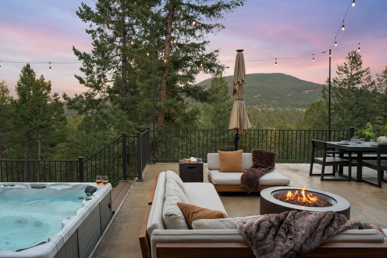 Outdoor terrace with a hot tub, fire pit, lounge seating, and forested mountain views at dusk.