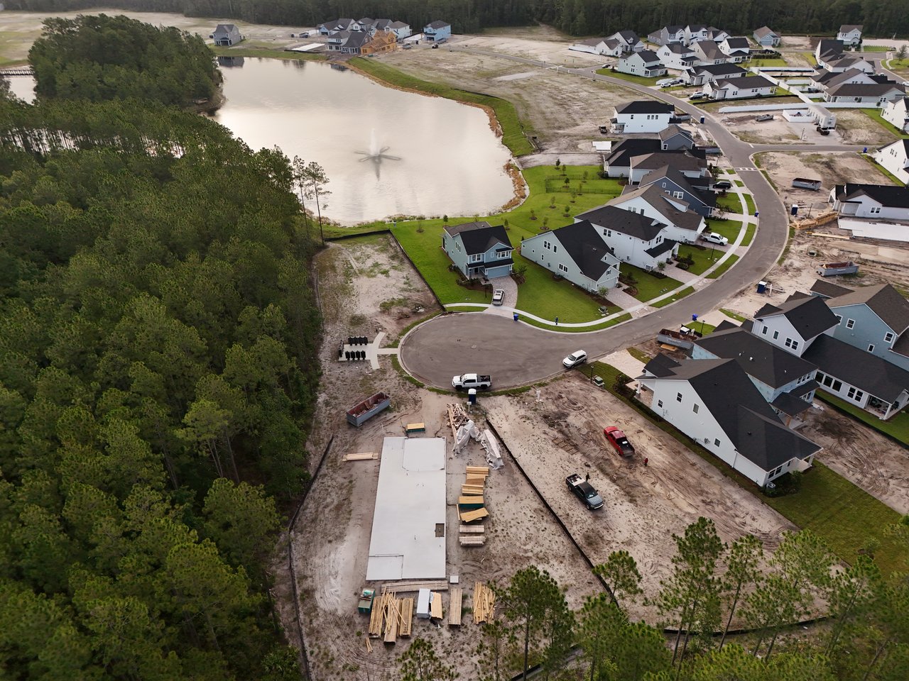 Aerial view of homes and construction sites around a lake beside dense forest.