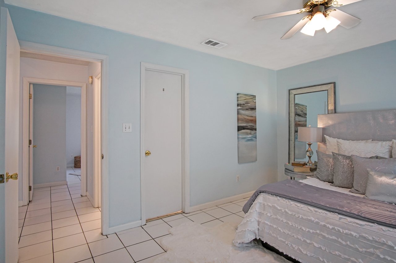 A serene bedroom with pastel blue walls, featuring a bed adorned with silver and white pillows. A ceiling fan, modern lamp, and abstract wall art enhance the decor.