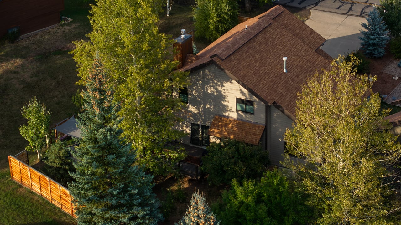 Tan house with brown shingle roof, trees, and wooden fence.