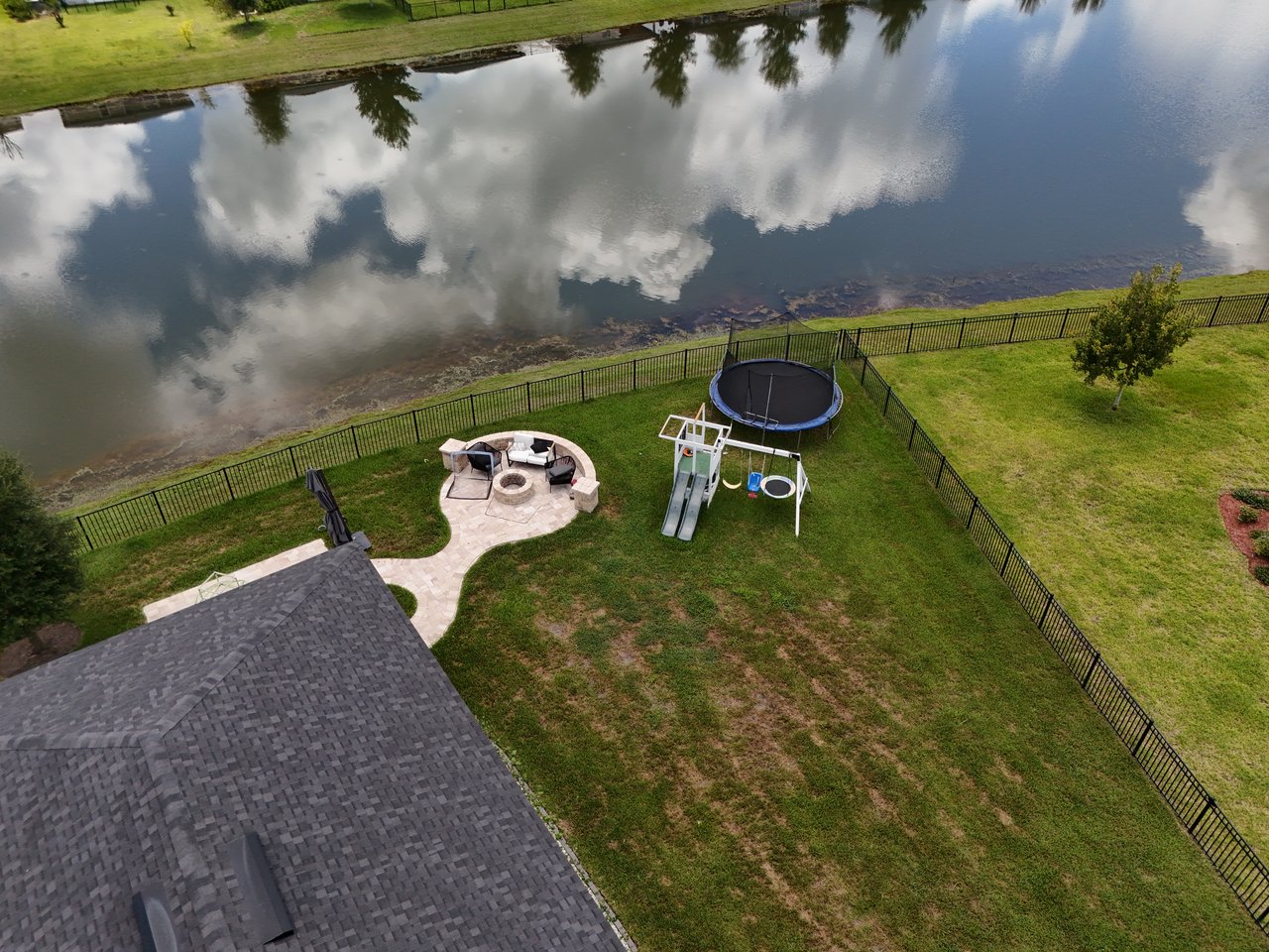 Fenced backyard beside a pond, with a fire pit patio, playset, and trampoline.