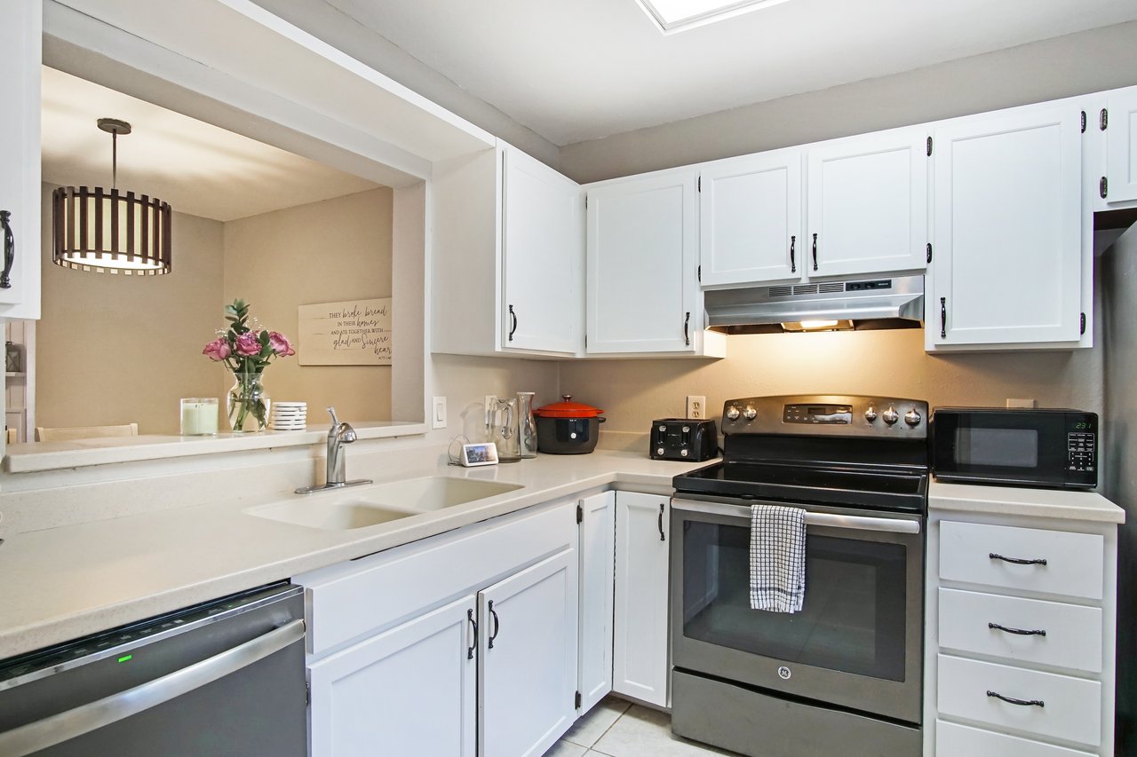 Modern kitchen with white cabinets, stainless steel appliances, and black oven. A countertop holds a red pot, toaster, and microwave, creating a cozy feel.