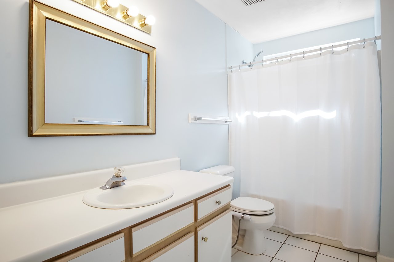 A bright, clean bathroom with a large mirror above a white sink. A shower with a white curtain is beside a toilet. The atmosphere is fresh and minimalistic.