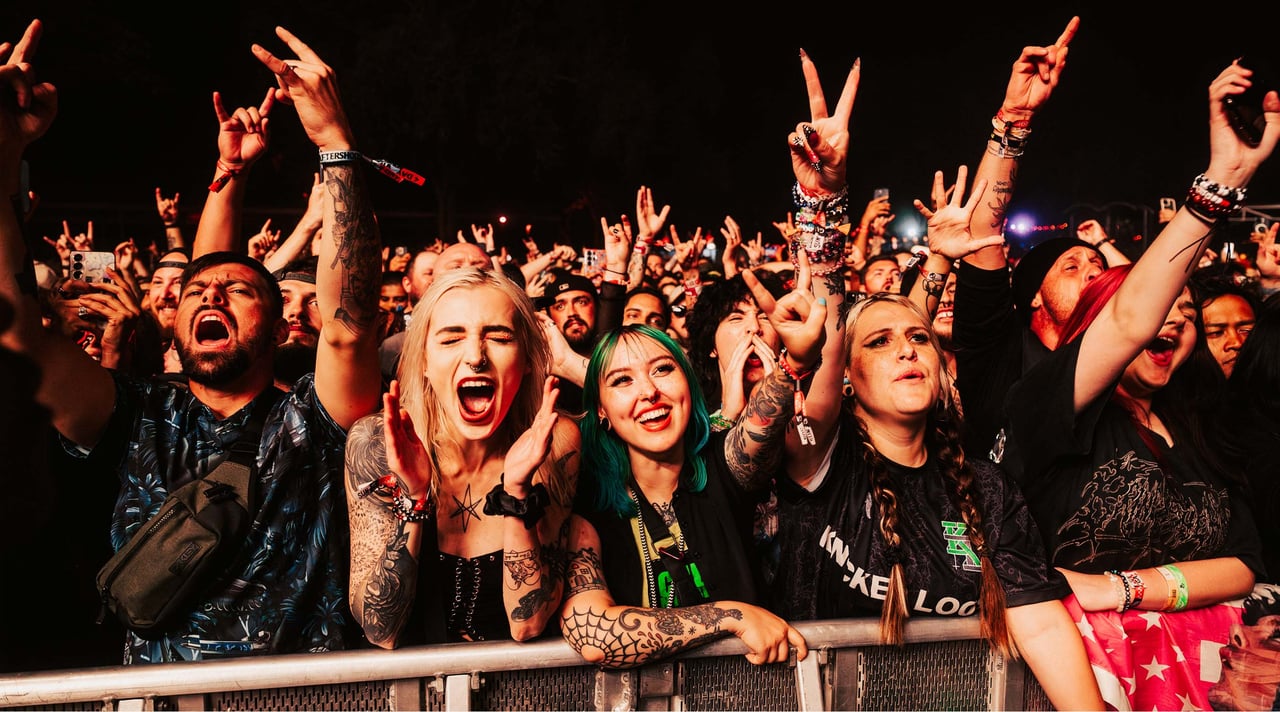 Concertgoers cheer and raise their hands behind a barrier, their faces lit by stage lights.
