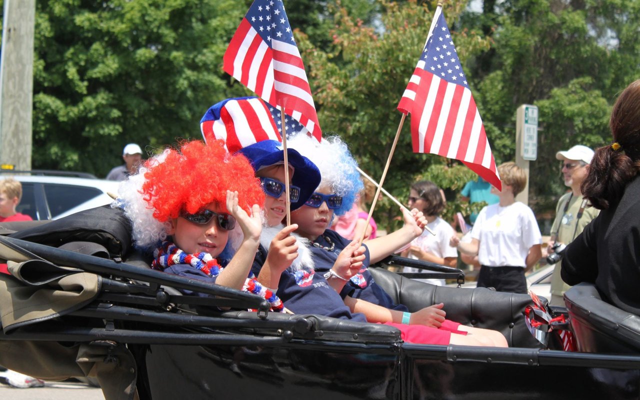 Children in patriotic wigs wave American flags from a parade vehicle.