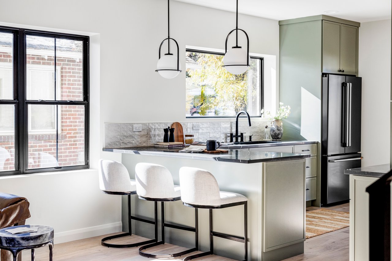 Airy and light modern kitchen with sage cabinets, black quartz countertops and fixtures, and three white barstools