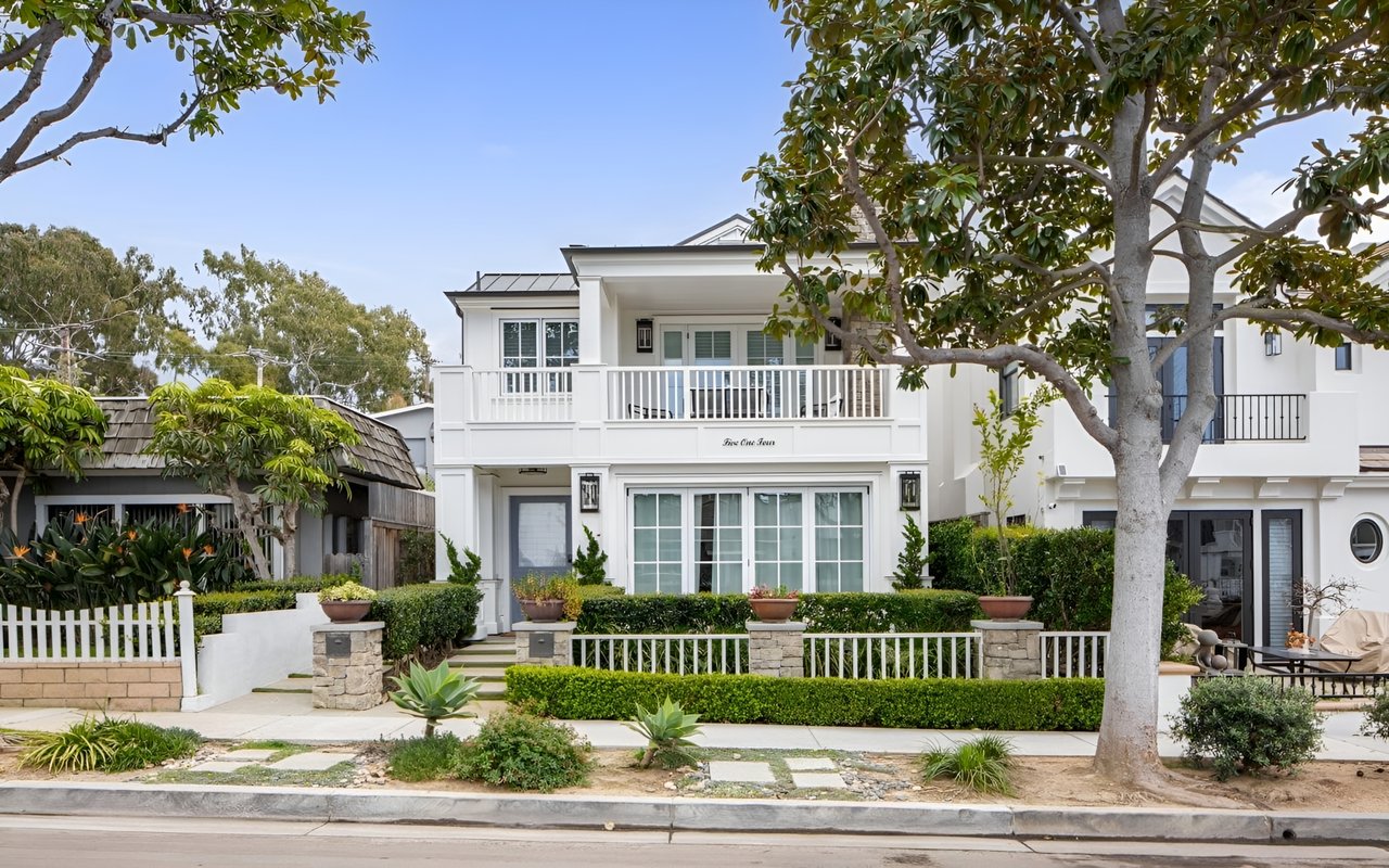  A large, modern, two-story white house with a balcony and a well-kept front yard with a white picket fence.