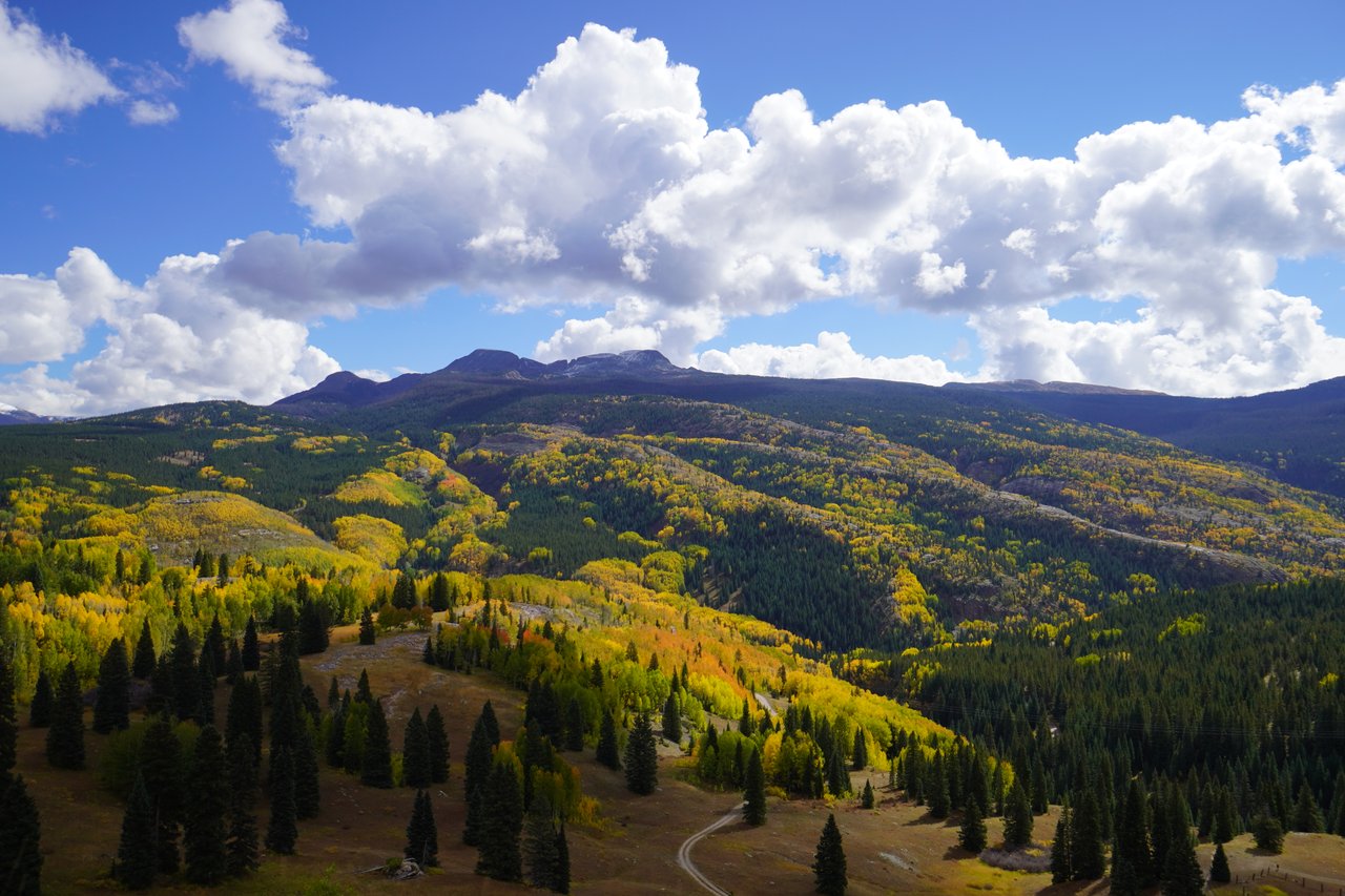 Autumn-colored forests cover mountain slopes beneath a bright blue sky with large white clouds.