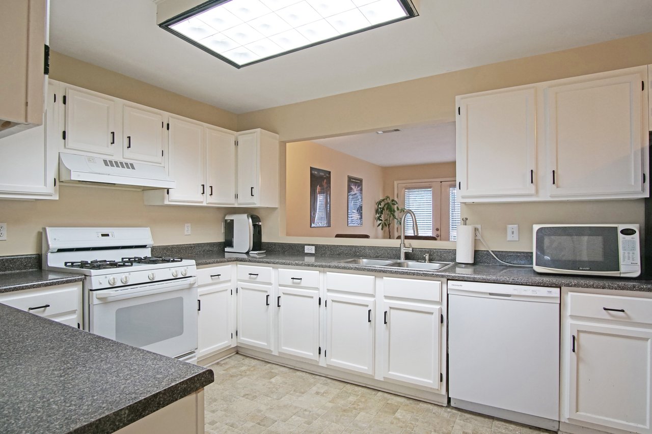 Kitchen at 529 Collinsford Road, Tallahassee, Florida, featuring white cabinetry with black hardware, gray countertops, and beige walls. The space includes a gas range, dishwasher, microwave, and double sink beneath a pass-through window that opens to the living area. A large ceiling light provides bright, even illumination.