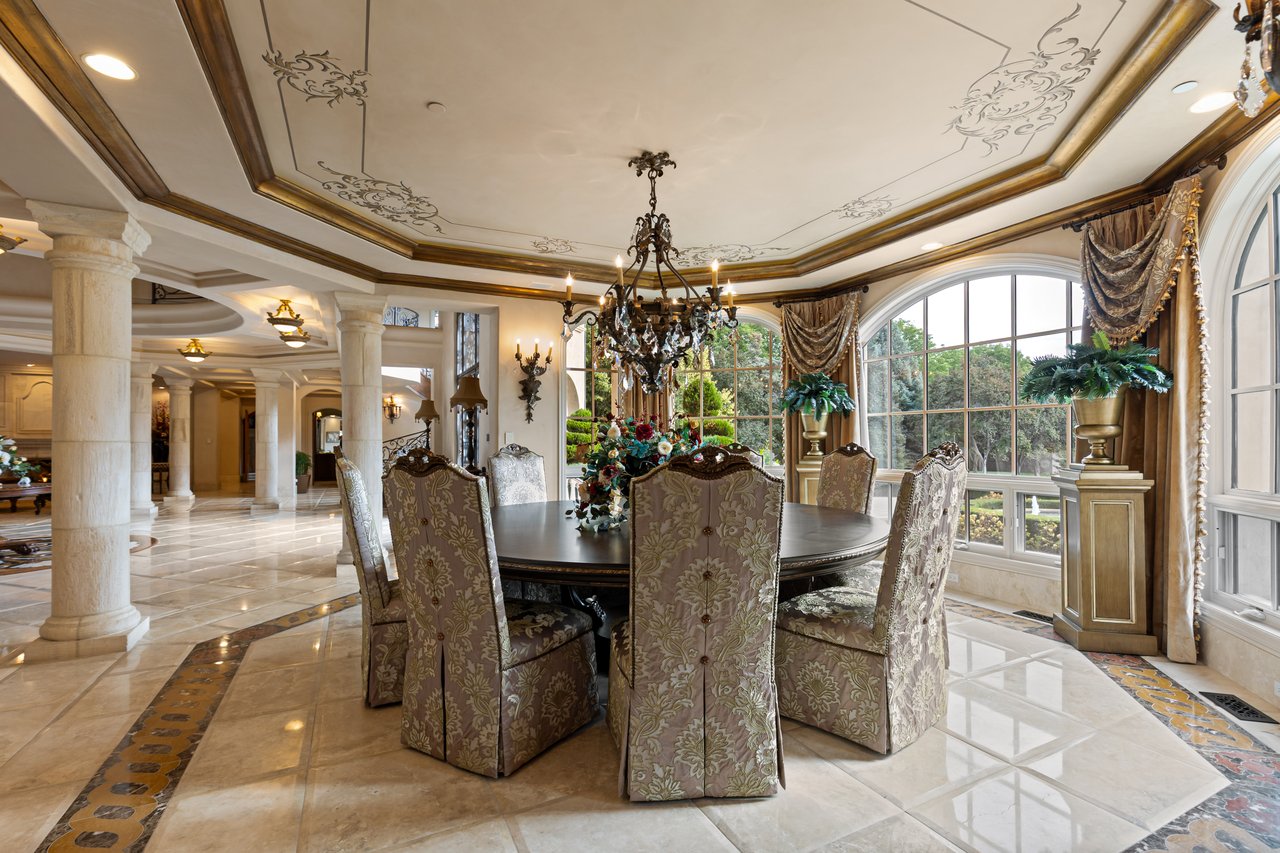 Dining room with an ornate ceiling, chandelier, round table, and tall arched windows overlooking greenery