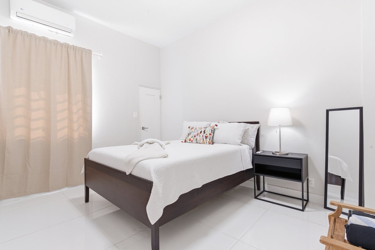 Furnished bedroom with a white-covered dark wood bed, beige curtains, and wall-mounted air conditioner.