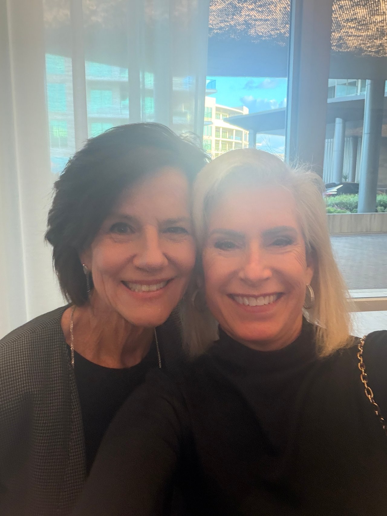Two smiling women pose together for a selfie by large windows.
