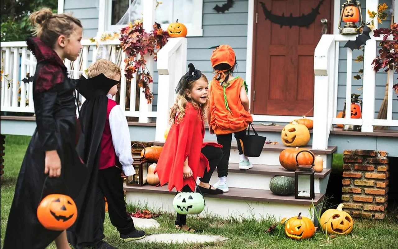 Children in Halloween costumes trick-or-treat at a pumpkin-decorated front porch.